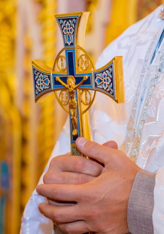 A priest in white vestments holding an ornate blue and gold cross