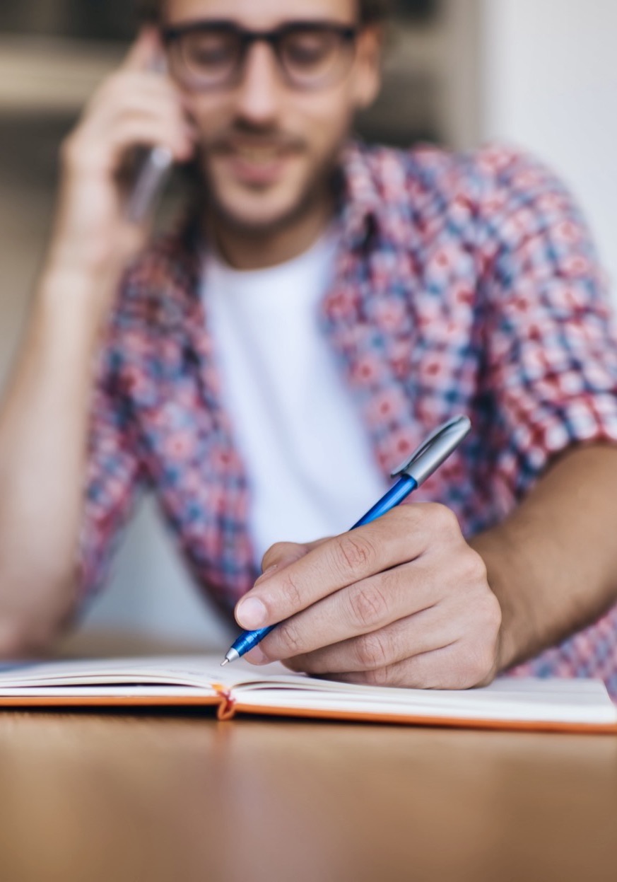 A parent on the phone writing notes beside an open laptop