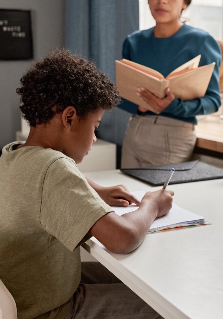 A student writing at a desk while an instructor reads aloud from a book