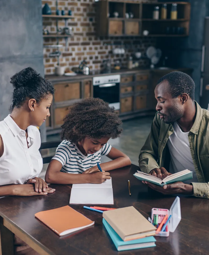 Young girl writing in a notebook at the kitchen table while her parents sit beside her