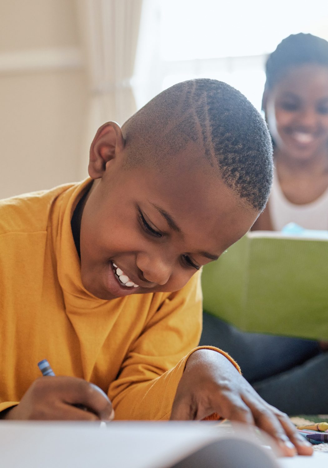 A smiling boy writing at a table while his mother reads behind him