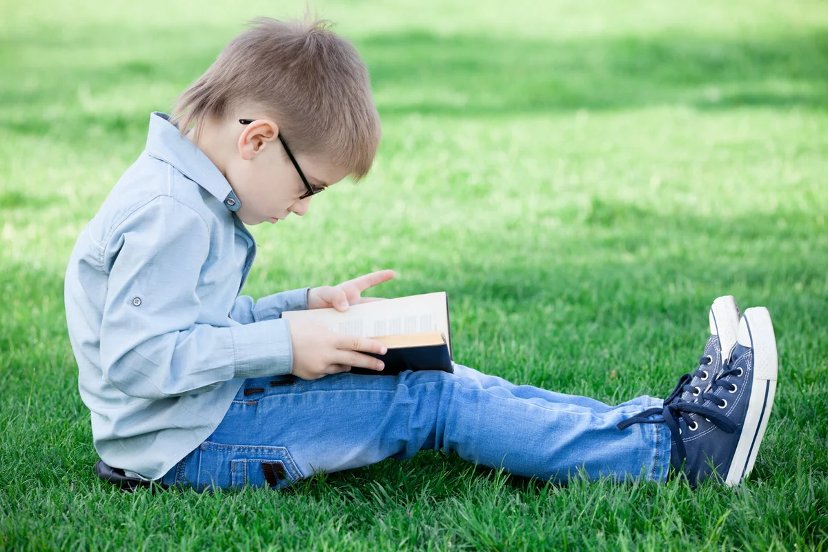 Young boy in glasses reading a book on the summer grass, a pick from a summer reading list for 3rd graders