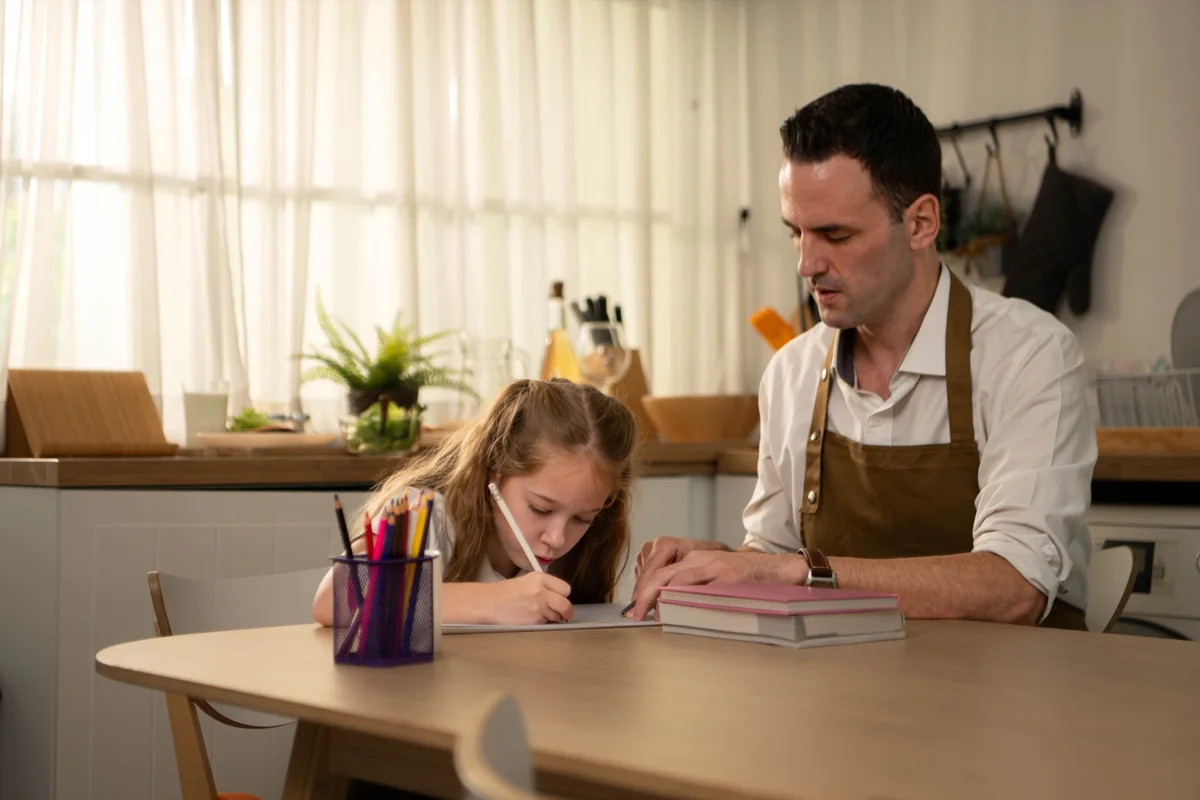 Father in an apron guiding his young daughter's writing at the kitchen table, classical education and learning differences