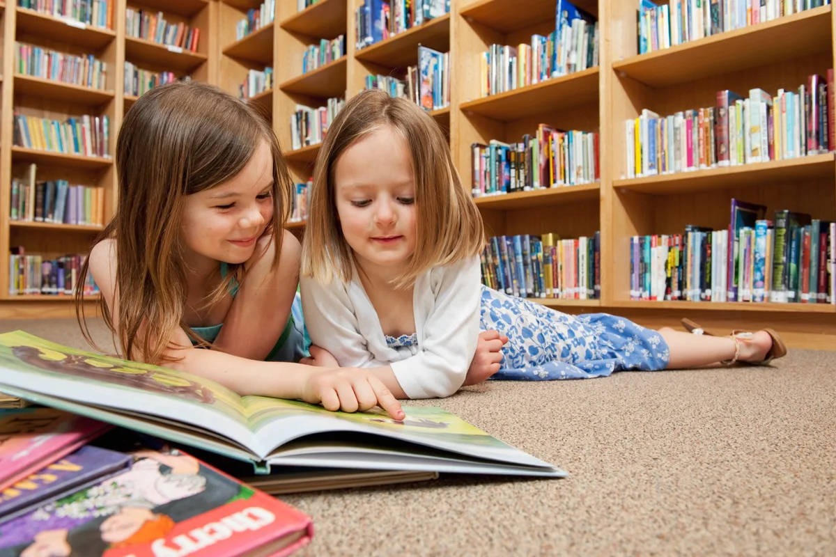 Two young girls reading a picture book together on a library floor, asking whether classical education is for everyone