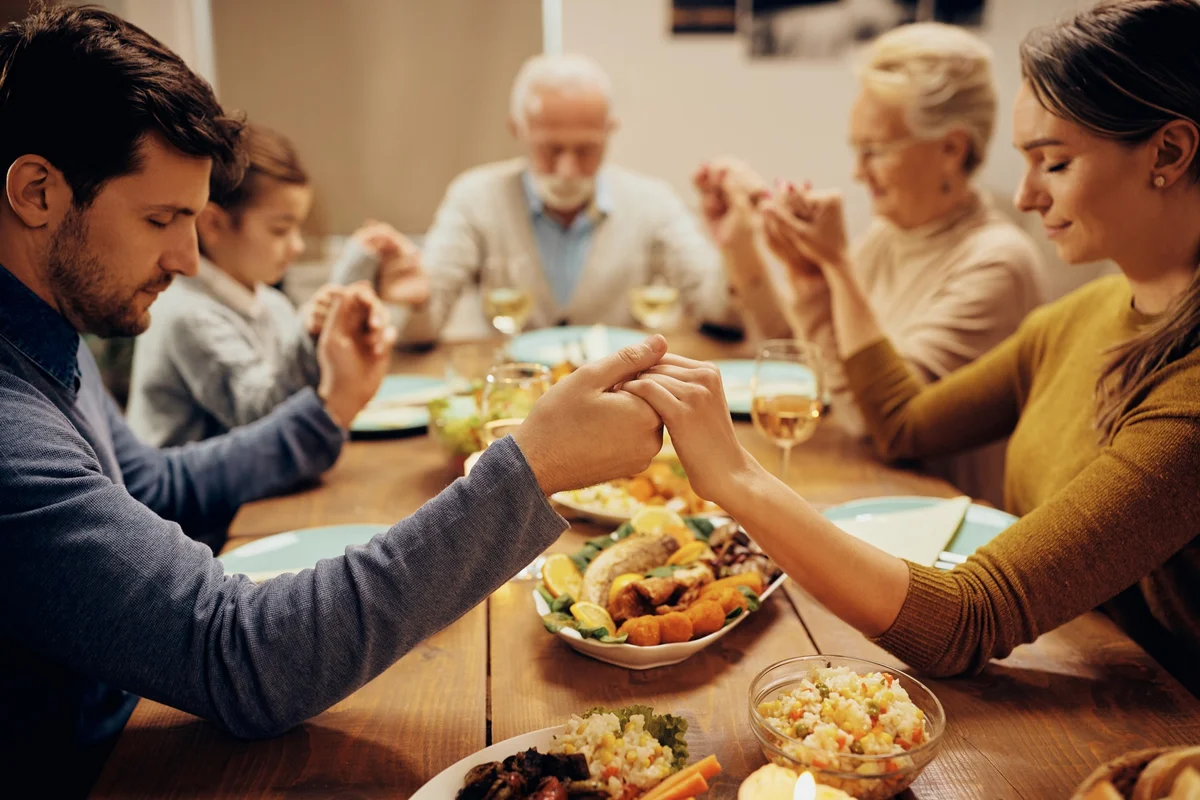 Three generations of a family joining hands in prayer around the dinner table, piety and justice taught at home