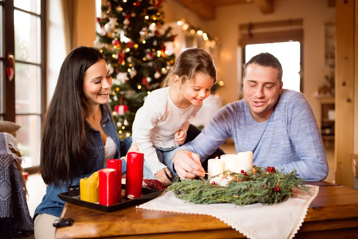 Parents and daughter lighting candles on a greenery wreath by the Christmas tree, passing down Christmas traditions