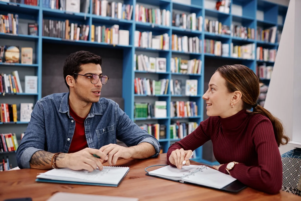 Two students talking over open notebooks in front of library shelves, a discussion rooted in the etymology of debate