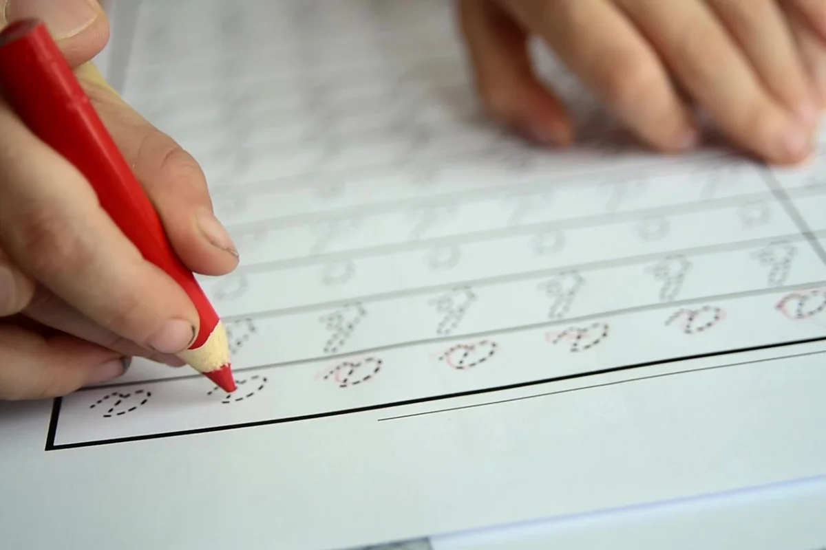 Child tracing a row of cursive letters with a red pencil, the kind of clear model rubrics in classical education provide