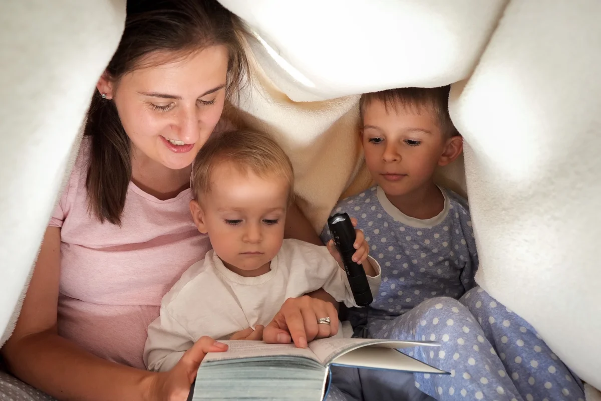 Mother and two small children reading by flashlight under a blanket fort, sharing middle grade read-alouds together