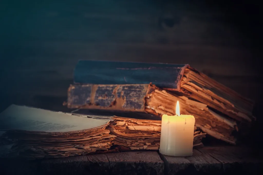 Candle lit beside old open books on a dark wooden table, the quiet start of a classroom liturgy