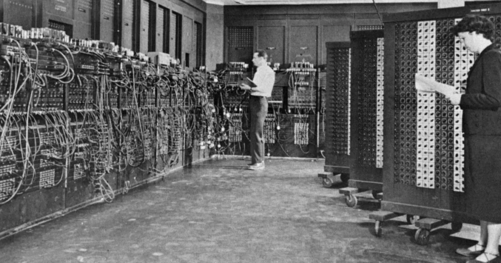 ENIAC computer room, a man and a woman reading notes beside walls of cable-covered panels and rolling function tables