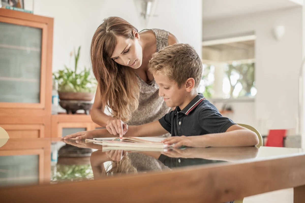 Mother leaning over her son at a dining table, pointing to a page in his schoolwork